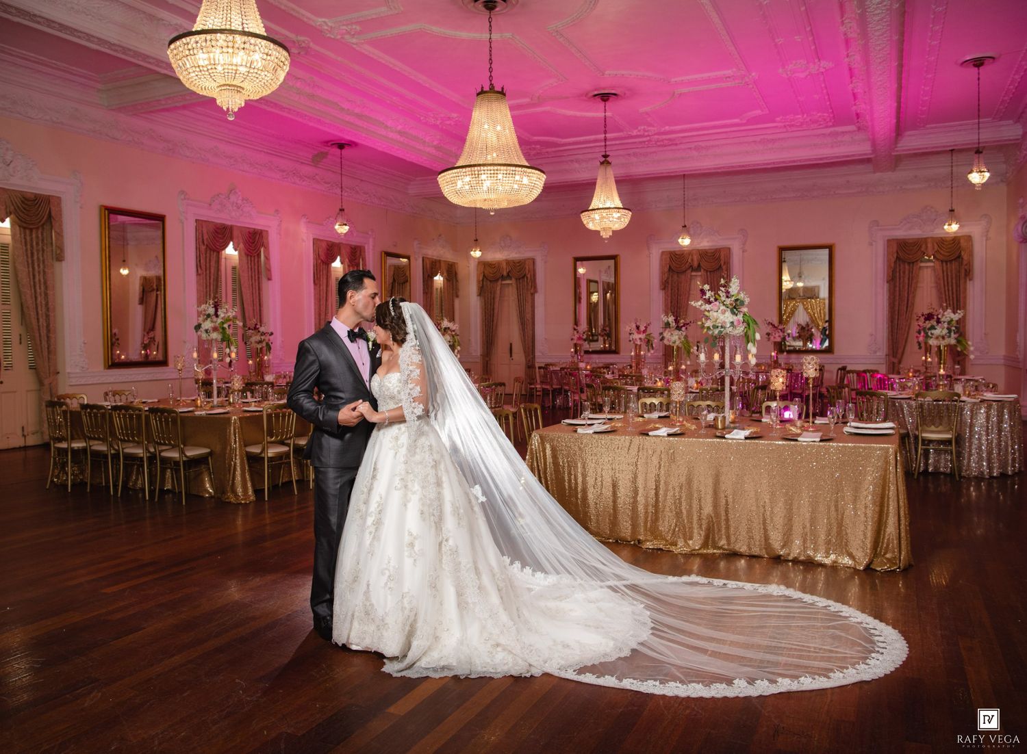 Bride and groom share a romantic dance in an elegant ballroom with chandeliers and pink uplighting.