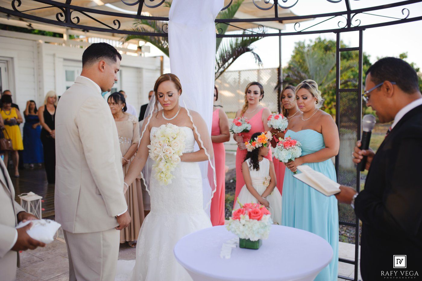 Bride and groom exchange vows at outdoor wedding ceremony with bridesmaids in teal dresses and floral arrangements.