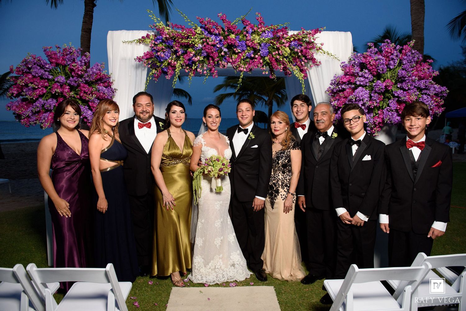 Wedding party poses under floral arch with purple bougainvillea at outdoor evening ceremony near ocean.