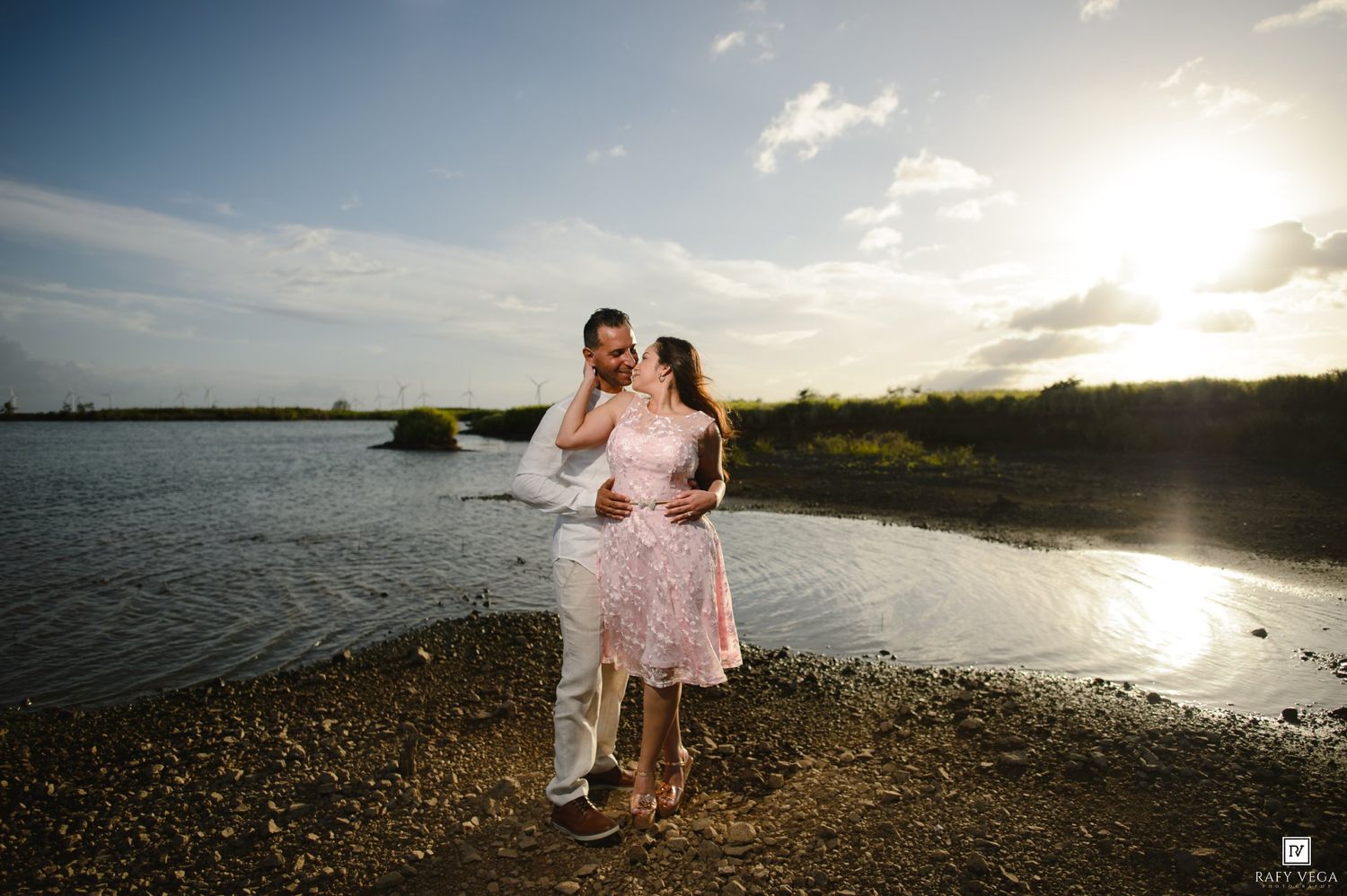 Couple embracing romantically by a serene lake at golden sunset, man in white shirt, woman in pink dress.