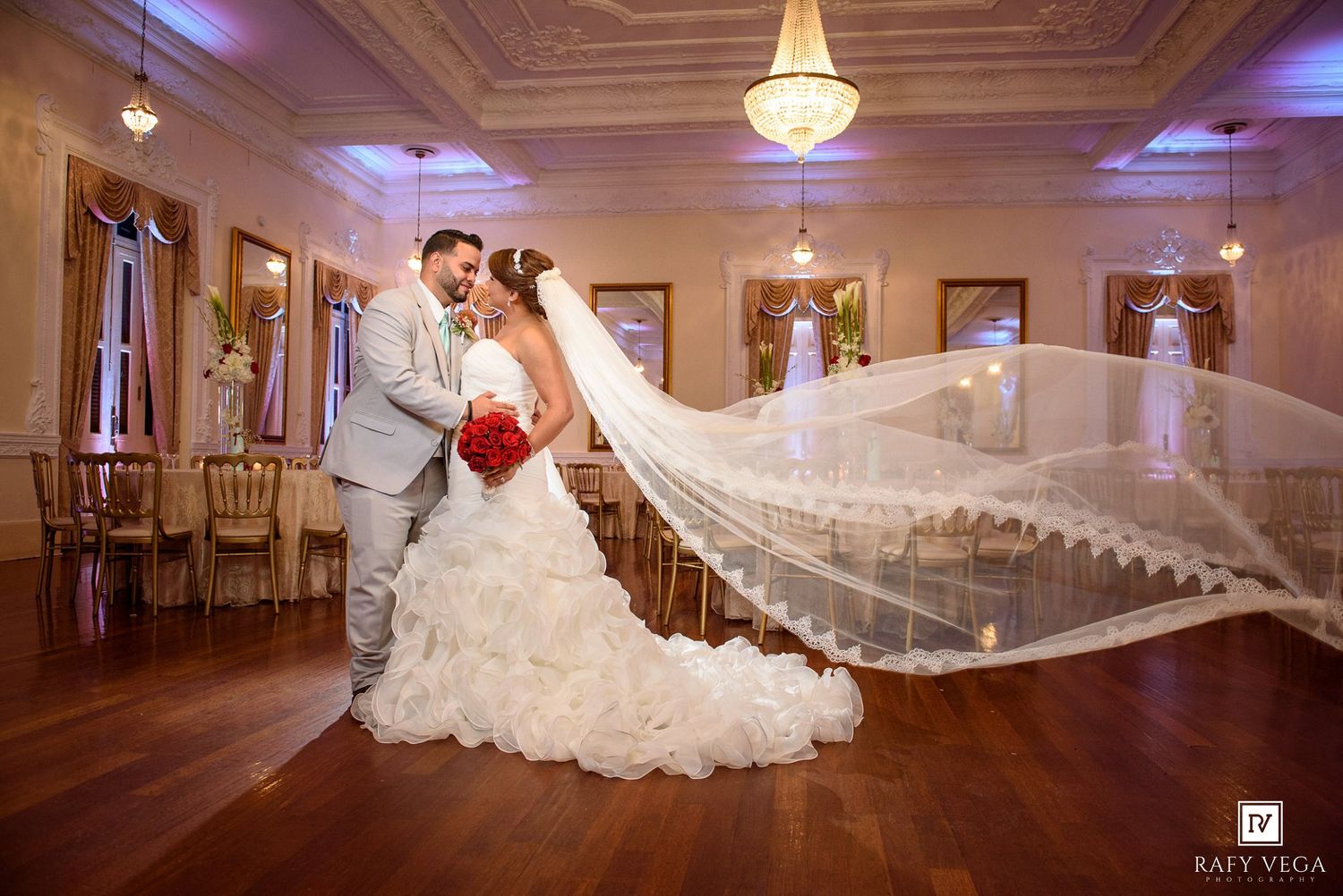 Bride and groom share romantic moment in elegant ballroom, bride's long veil flowing dramatically behind her.