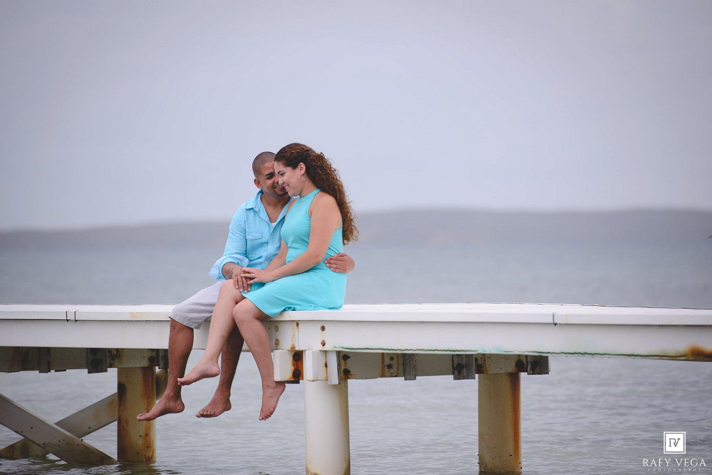 Couple in matching turquoise outfits sitting together on a seaside pier, sharing a romantic moment by the ocean.