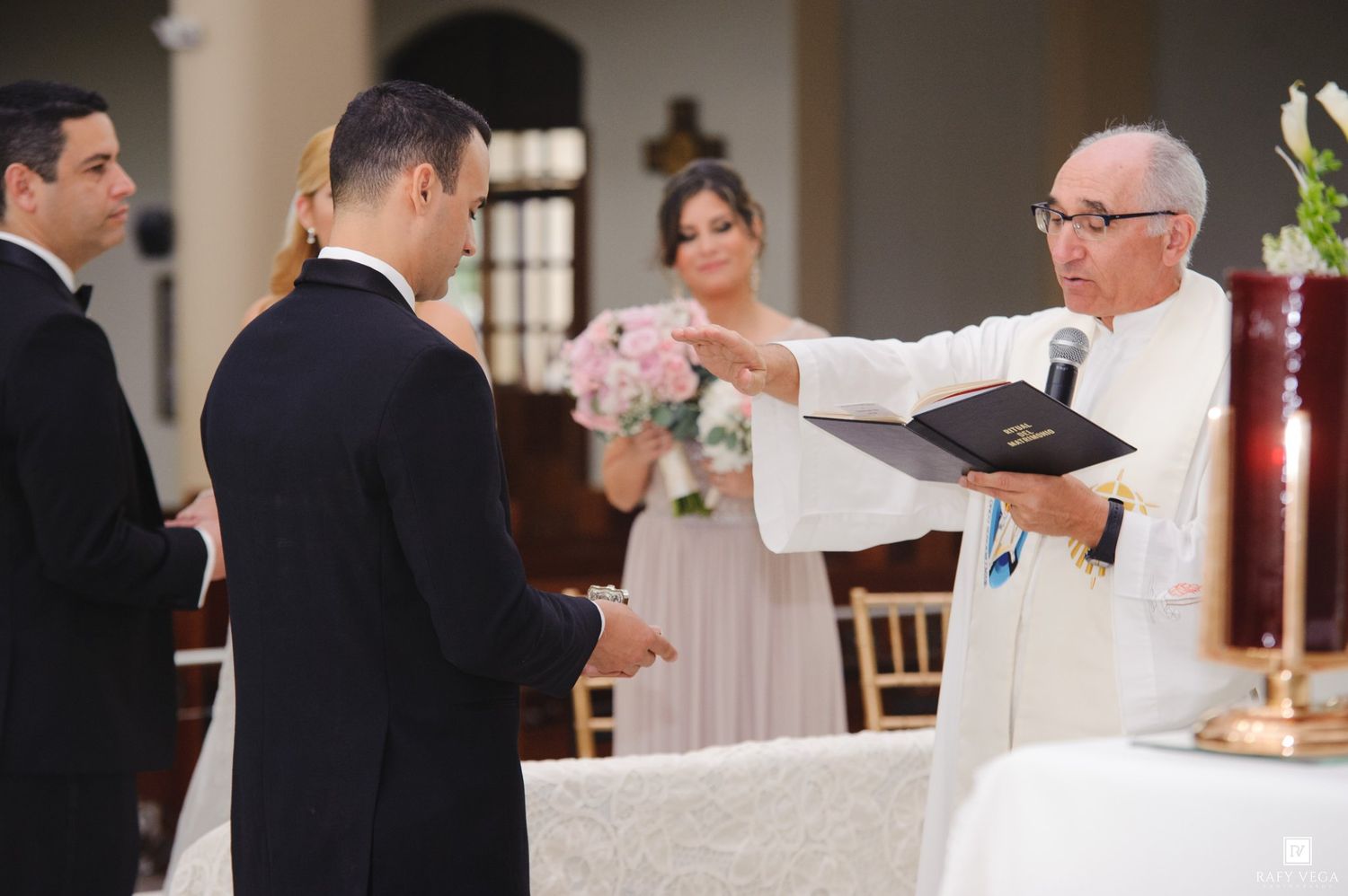Priest officiating a wedding ceremony, reading from a book, with groom and guests in a decorated church.