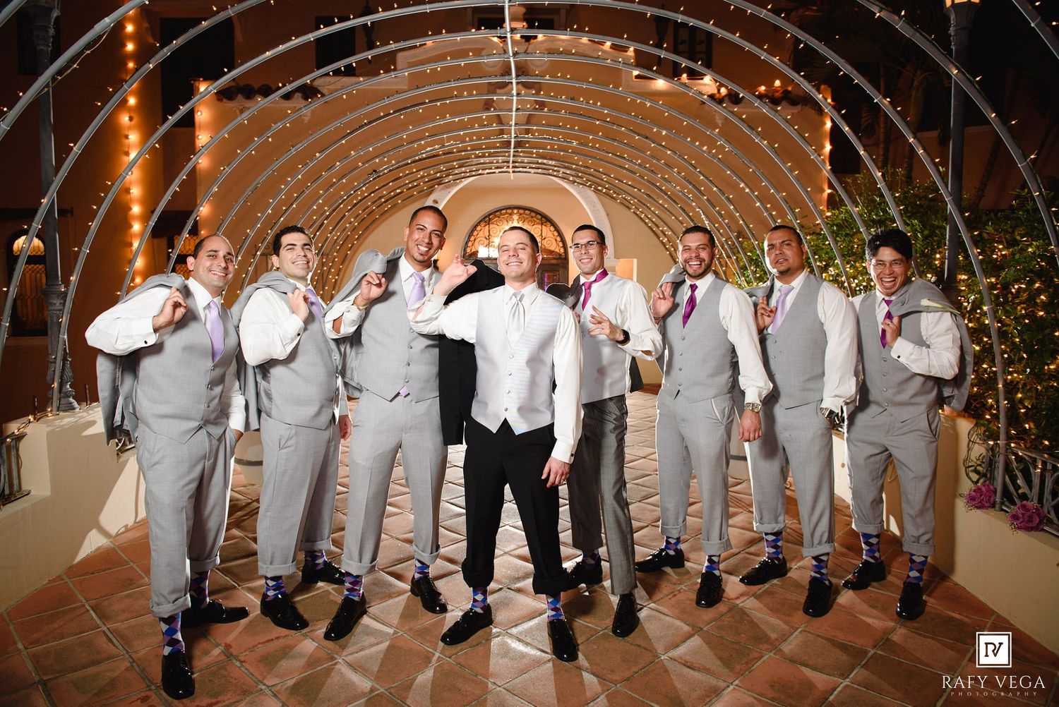Groom and groomsmen in gray suits pose playfully under string lights at an elegant wedding venue.