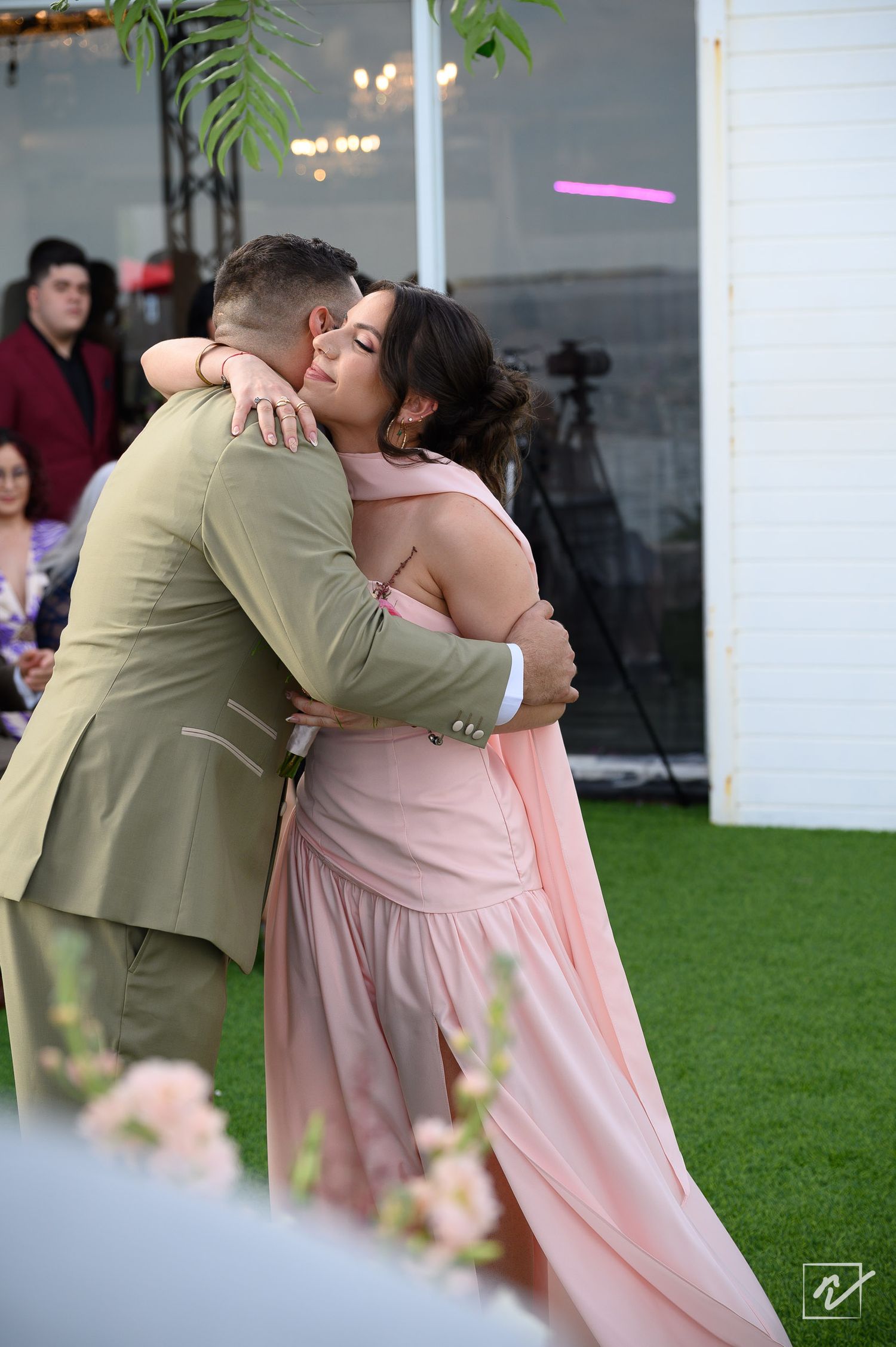 A woman in a blush pink dress and a man in an olive suit share a warm embrace at an outdoor wedding reception on a green lawn.