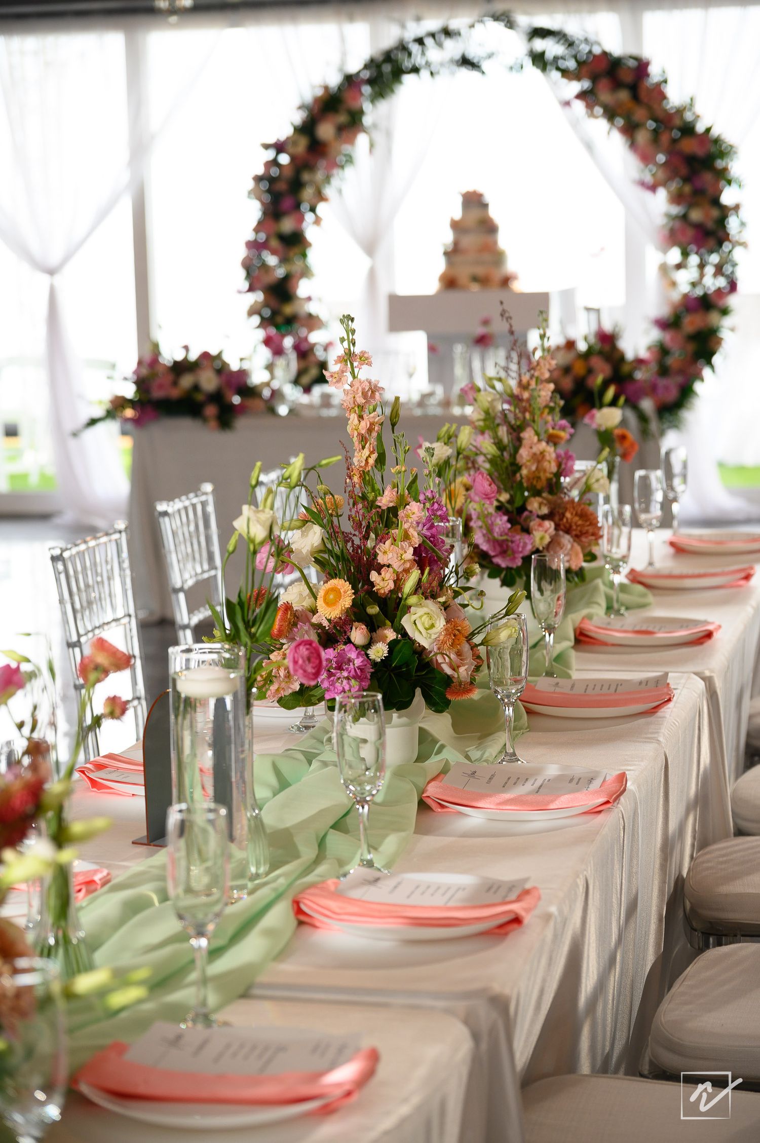 Elegant wedding reception table with colorful floral centerpieces, coral napkins, green runner and a floral arch beside a cake.
