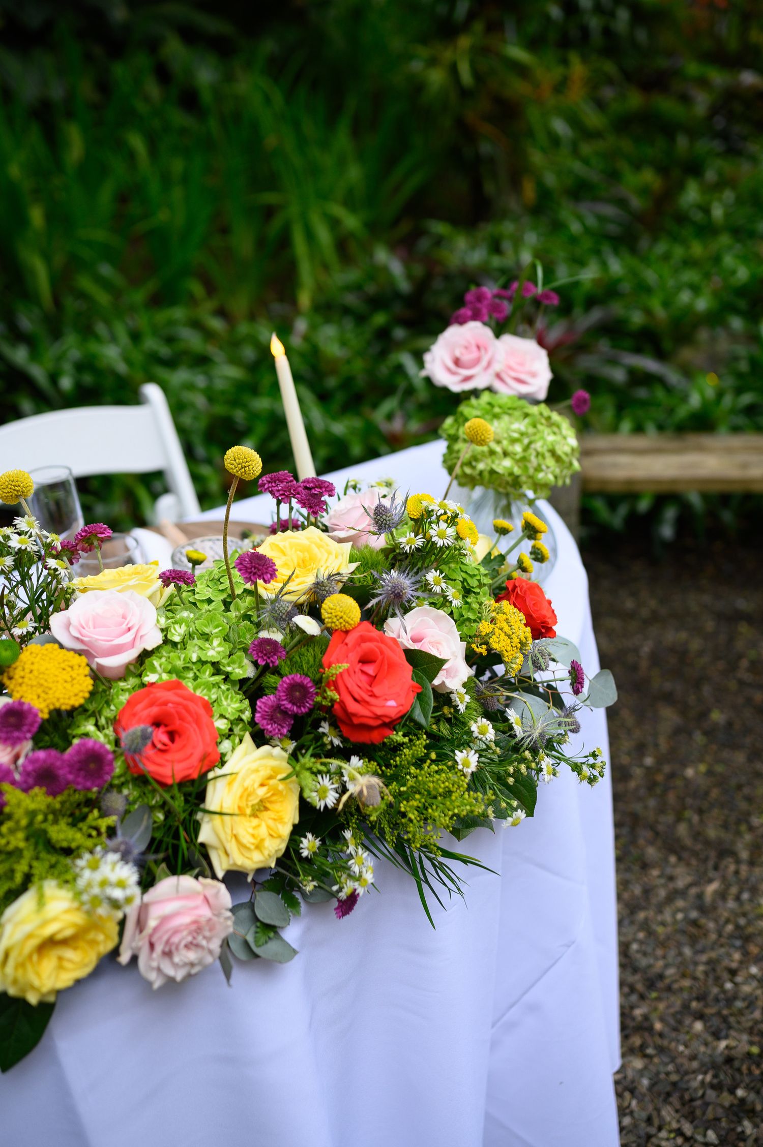 Colorful floral centerpiece with roses, yellow blooms, and greenery on a white tablecloth at an outdoor garden event.