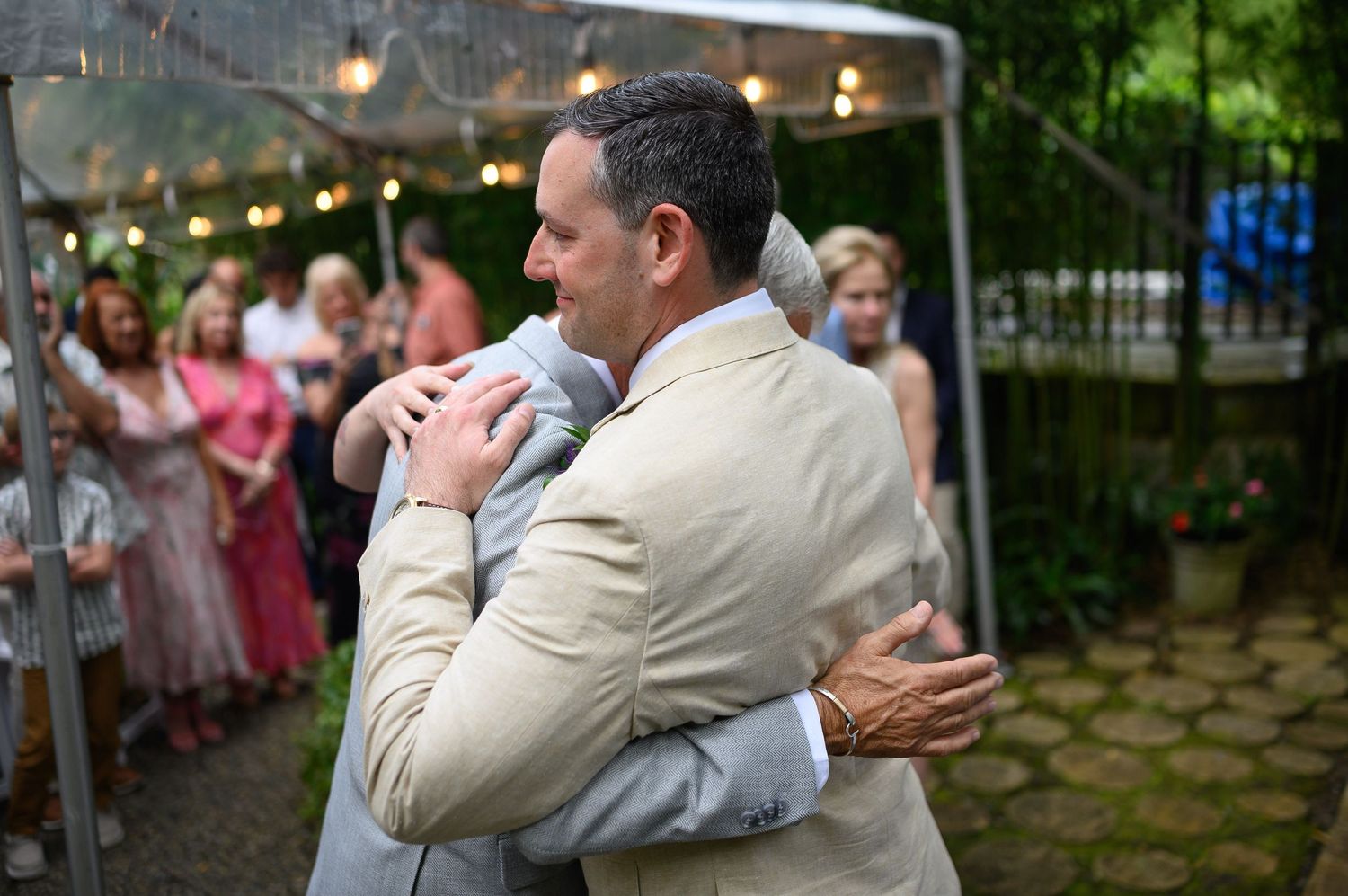 Two men share a heartfelt hug at an outdoor wedding reception with guests in background.