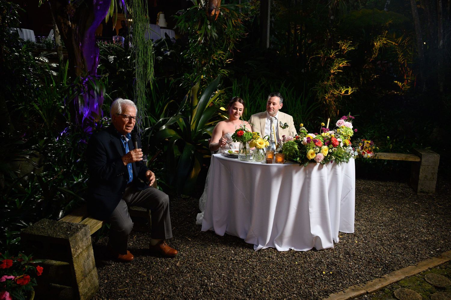 Elderly man speaks at outdoor wedding reception as bride and groom sit at floral-decorated sweetheart table at night.