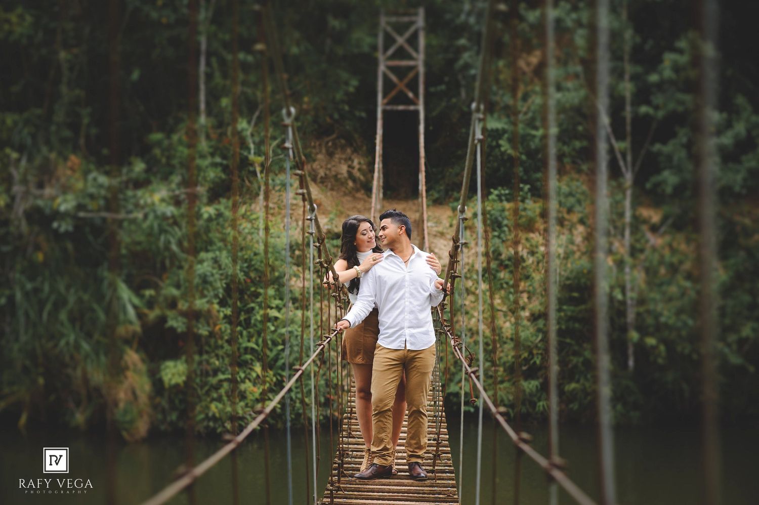 Couple embracing on a rope suspension bridge surrounded by lush green forest and a river below.