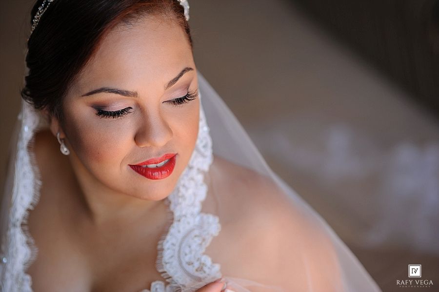Elegant bride in white lace veil and red lipstick, looking down gracefully in a stunning bridal portrait.