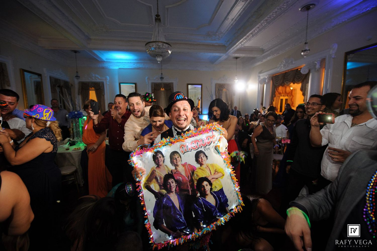 People dancing at a festive party, man holding colorful poster with Mardi Gras beads in decorated ballroom.
