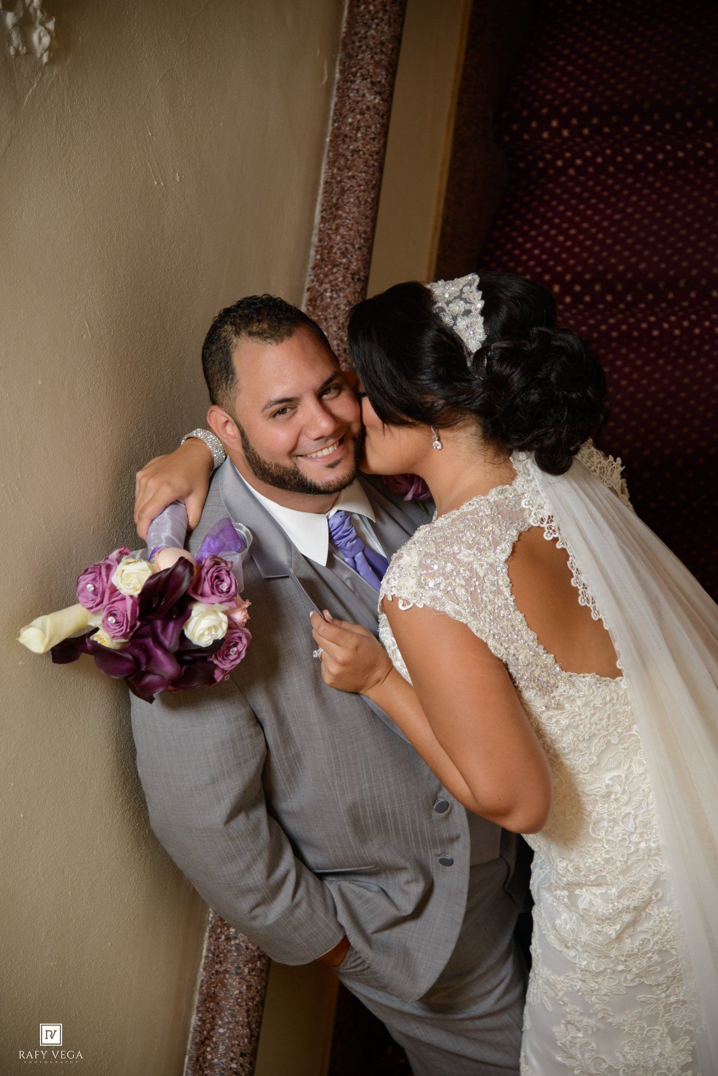 Bride in lace wedding gown kisses groom in gray suit holding purple bouquet during romantic wedding portrait.