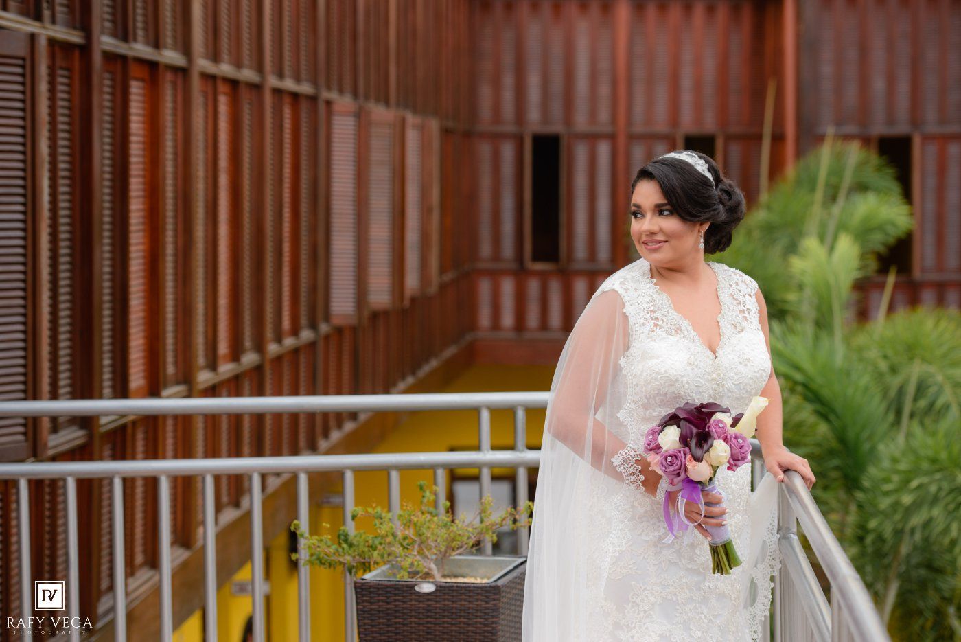 Bride in white lace gown holding purple bouquet poses on balcony with wooden architecture backdrop.