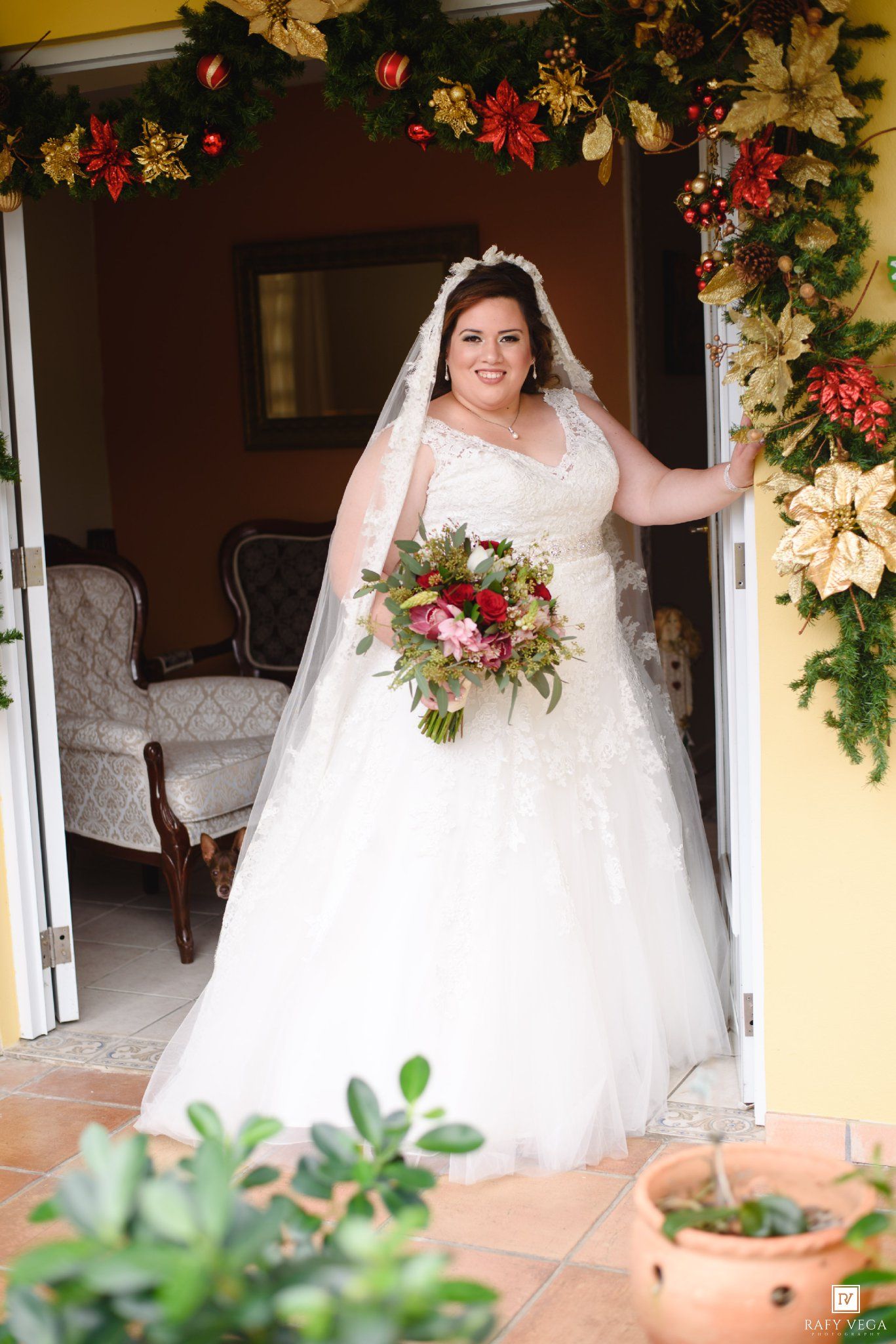 Bride in a white lace ball gown with veil holding a bouquet, standing in a Christmas-decorated doorway.