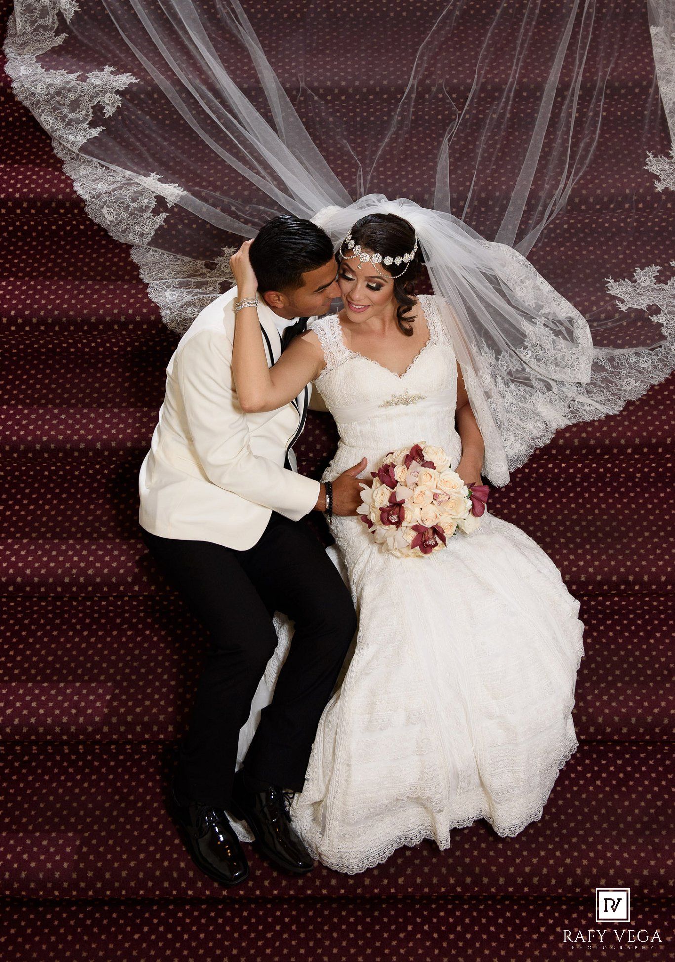 Bride and groom share a romantic kiss on red carpeted stairs, her lace veil flowing gracefully above them.