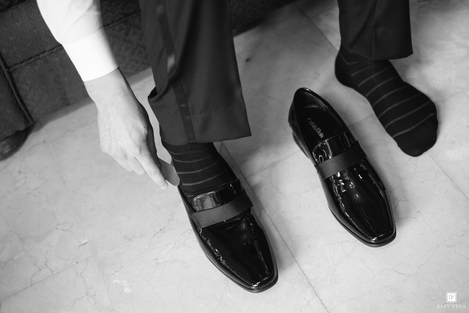 Black and white photo of a man putting on shiny patent leather loafers with dark socks and dress trousers.