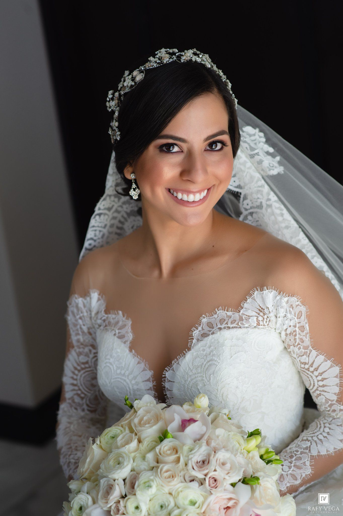 Smiling bride in lace wedding dress with veil and crystal headpiece holding white rose bouquet.