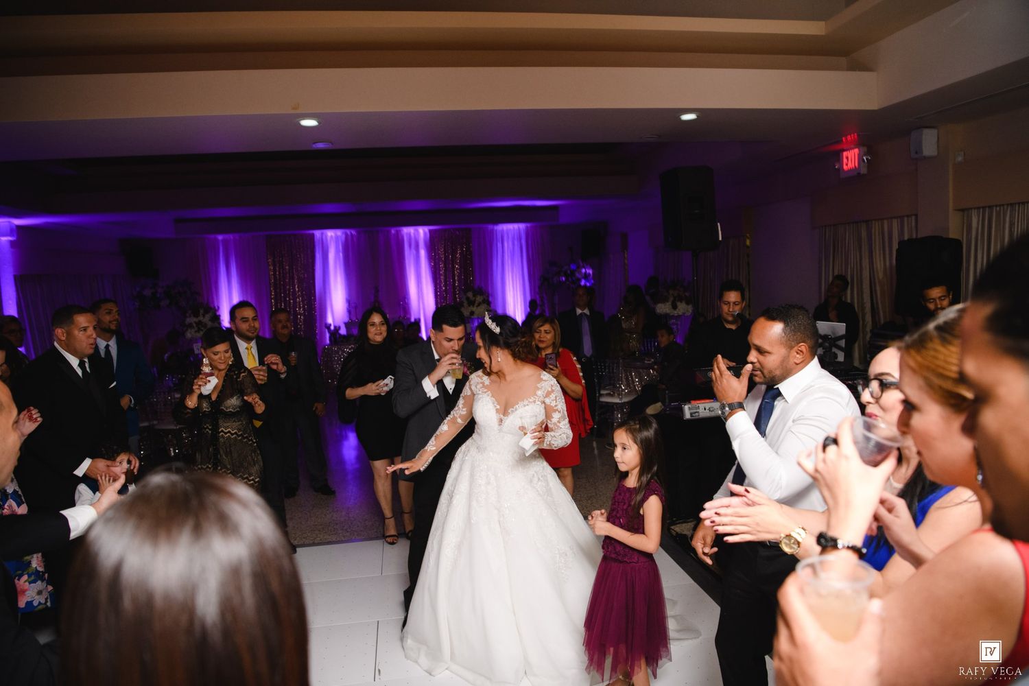 Bride and groom share first dance surrounded by cheering guests at elegant wedding reception with purple uplighting.