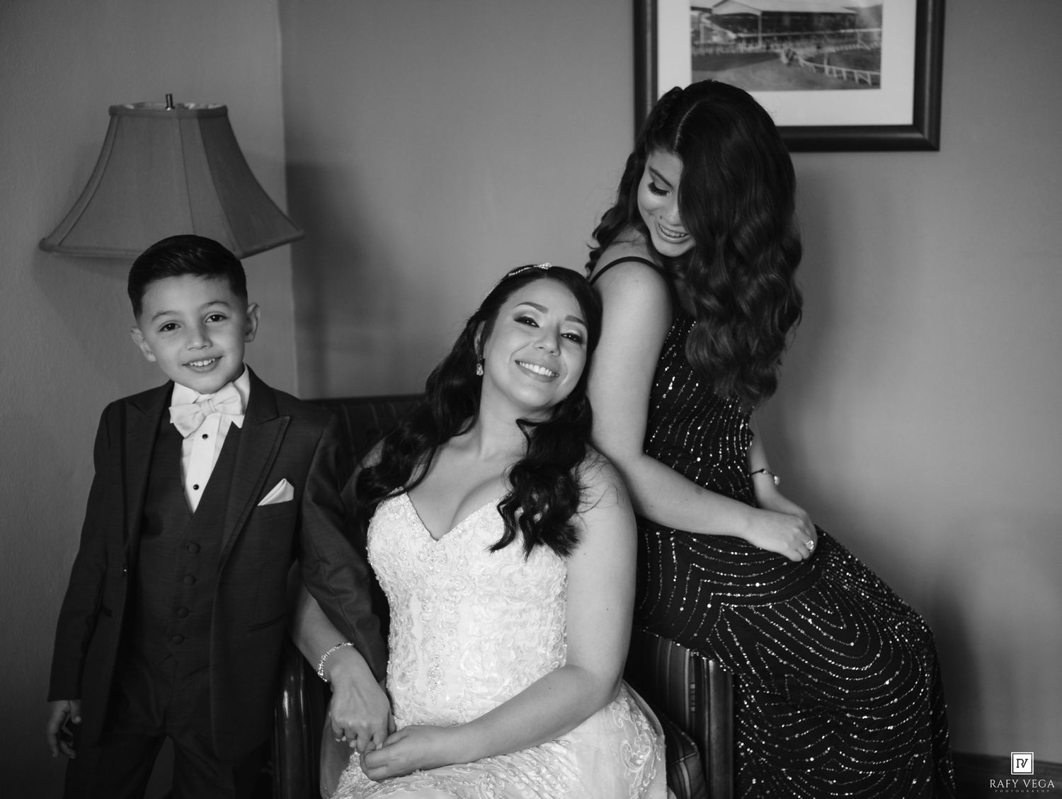 Black and white photo of a bride in a white gown with a young boy in a suit and a woman in a sparkly dress.
