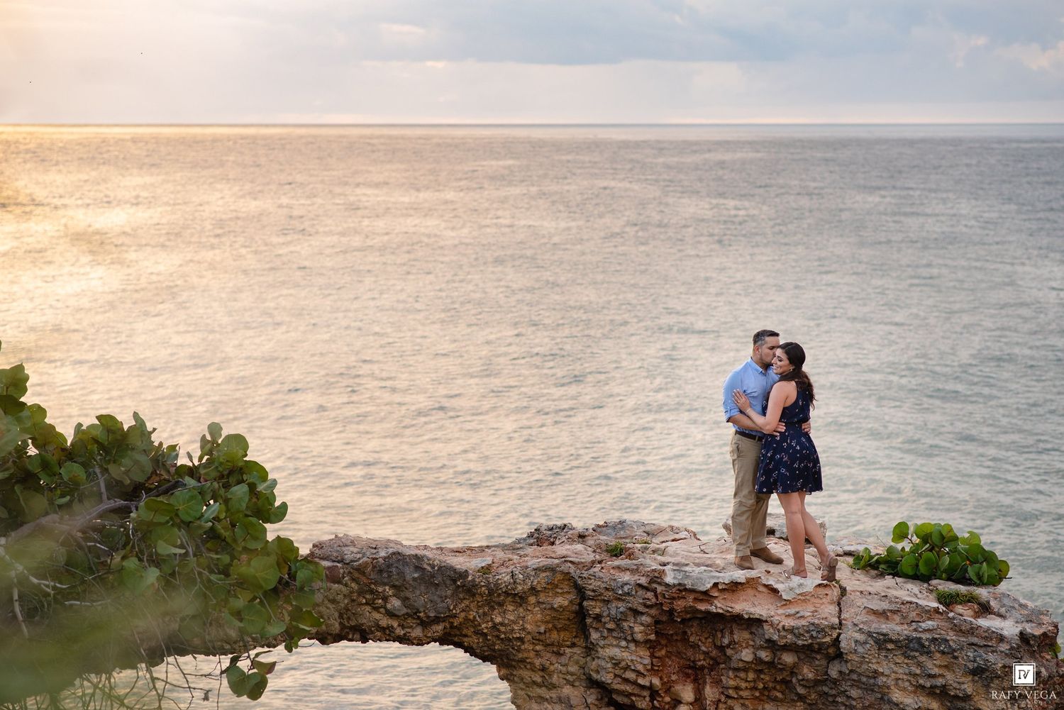 Couple embracing on rocky ocean cliff at sunset, with turquoise water and golden sky in the background.