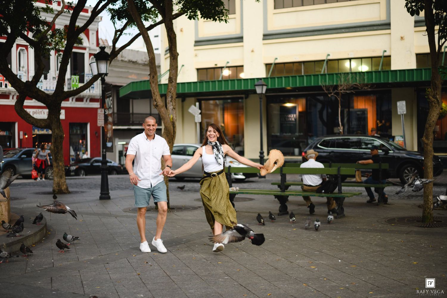 Couple joyfully holding hands and running through a city plaza surrounded by pigeons and green storefronts.