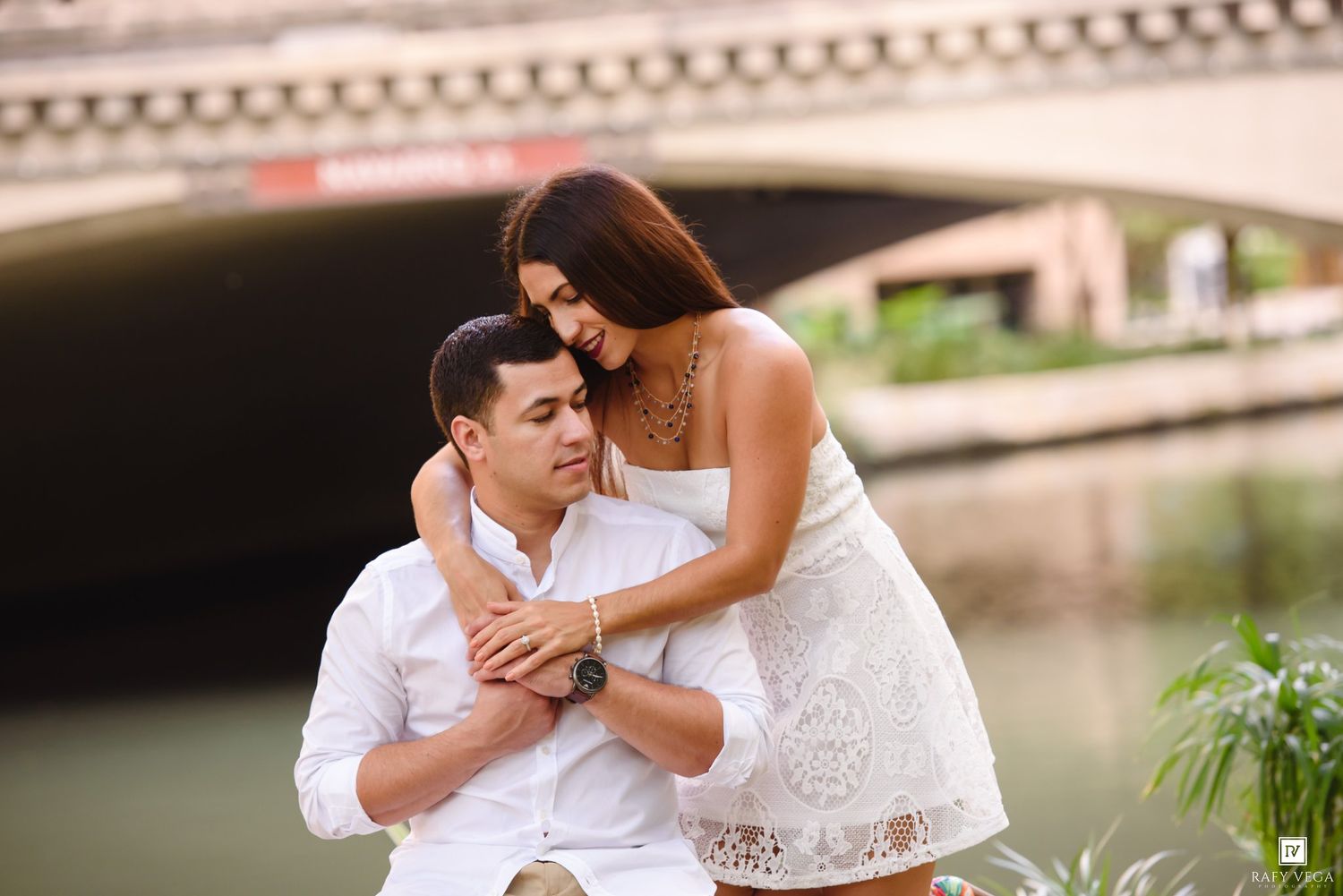 Couple in white outfits embracing romantically near a river with an arched bridge in the background.
