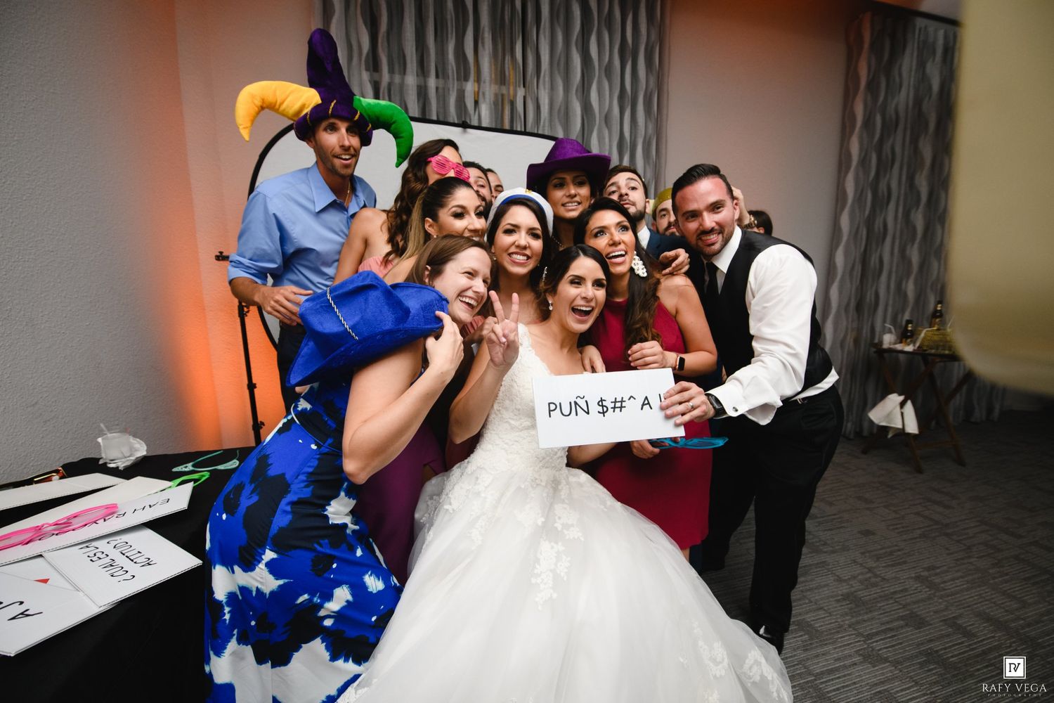 A bride in a white gown celebrates with wedding guests using fun photo booth props at a lively reception.