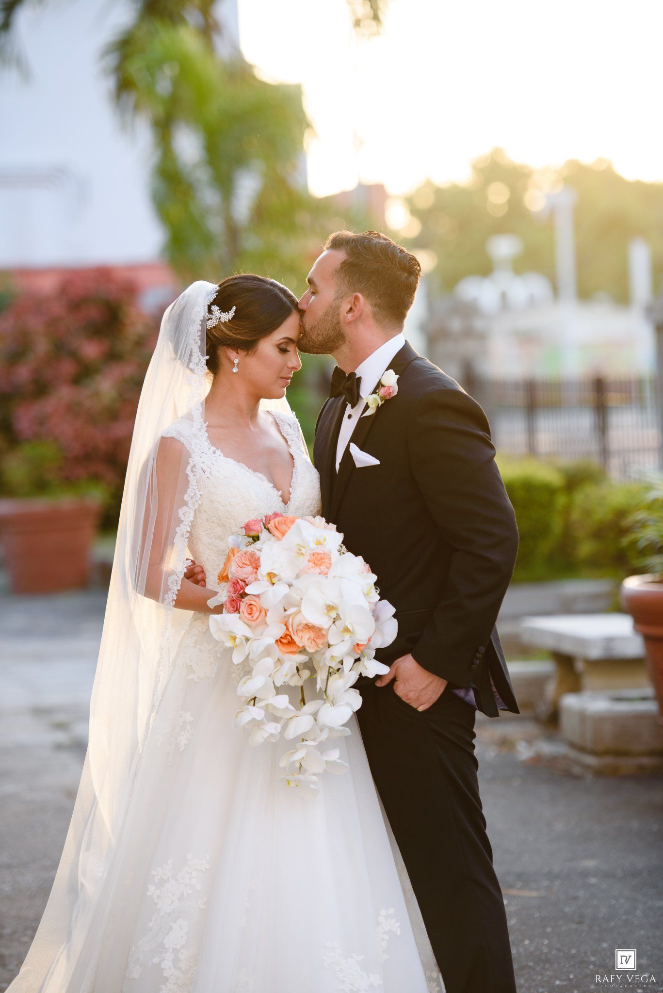 Bride in white gown with veil holds floral bouquet as groom in black tuxedo kisses her forehead outdoors.
