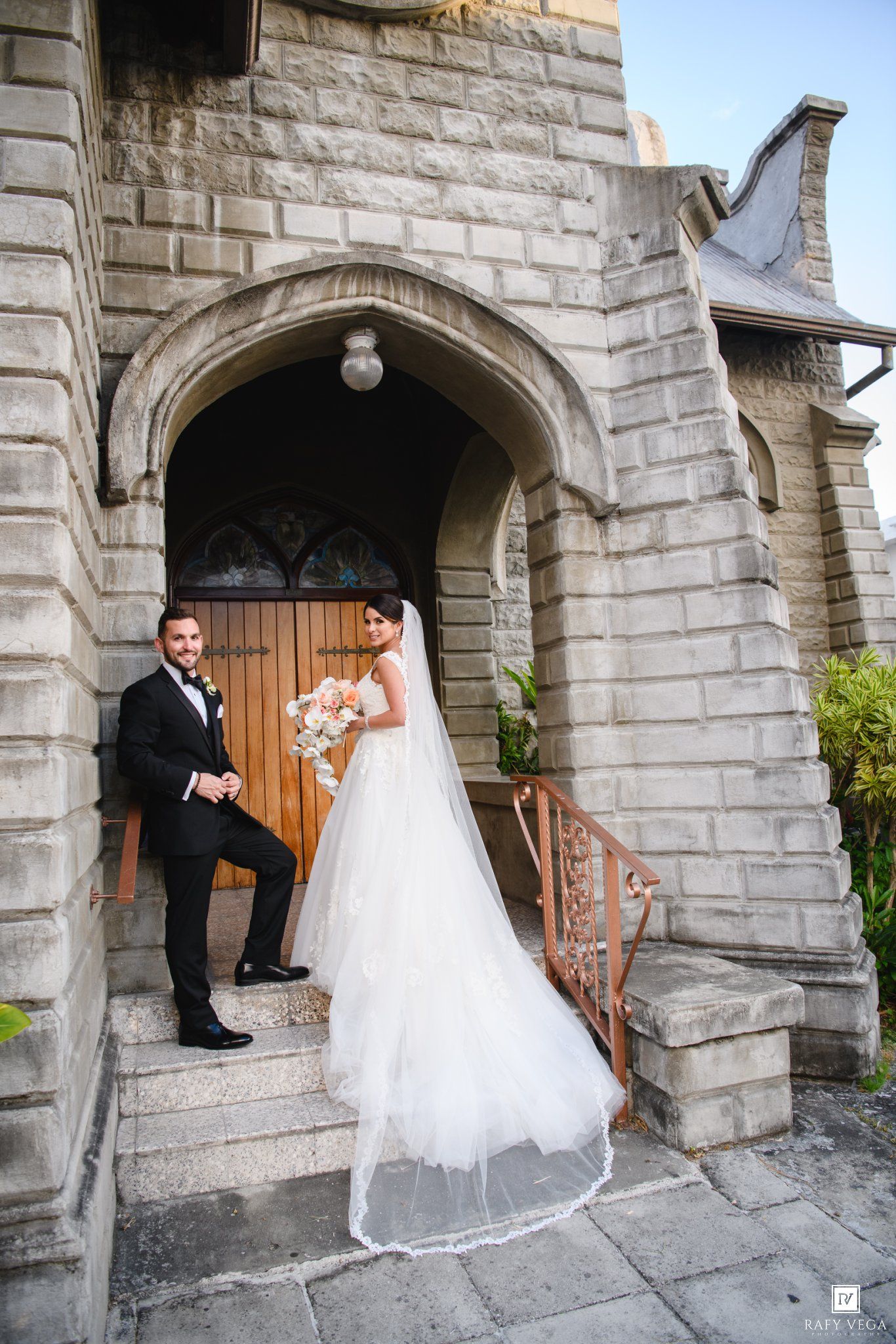 Bride and groom posing on stone church steps, bride in white gown with veil, groom in black tuxedo, holding bouquet.