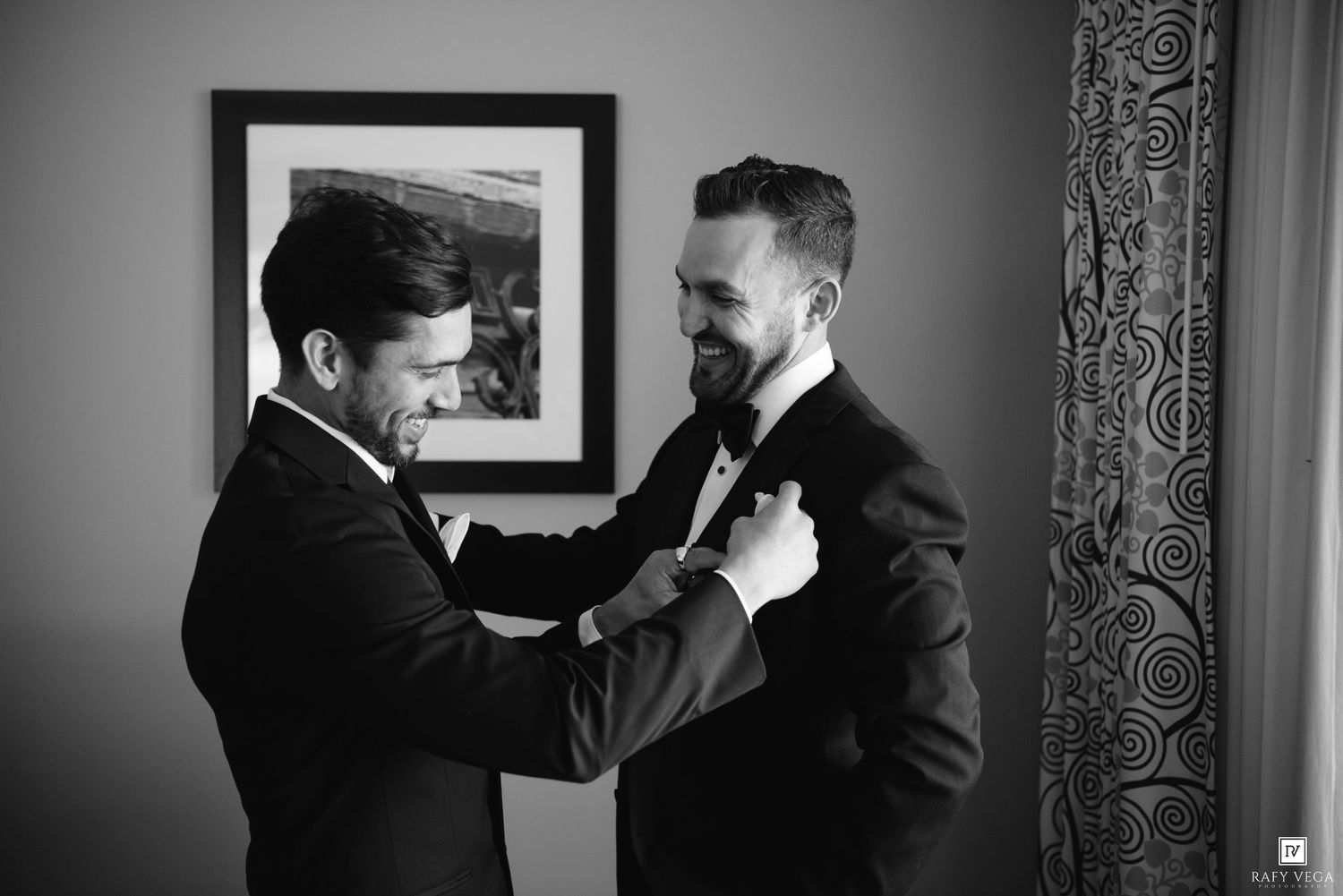 Two grooms in tuxedos help each other get ready in a black and white wedding preparation photo.