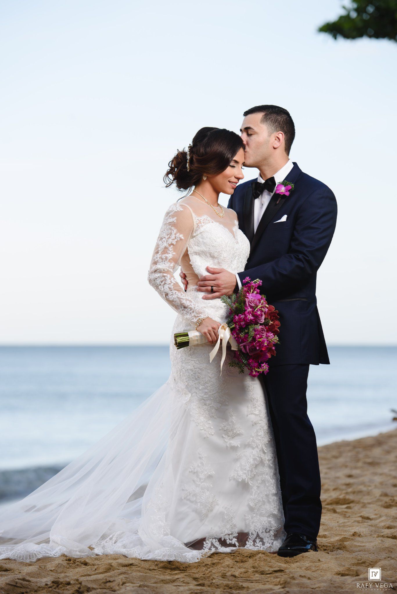 Bride and groom share a romantic kiss on a beach, bride in lace gown holding pink bouquet, groom in navy suit.