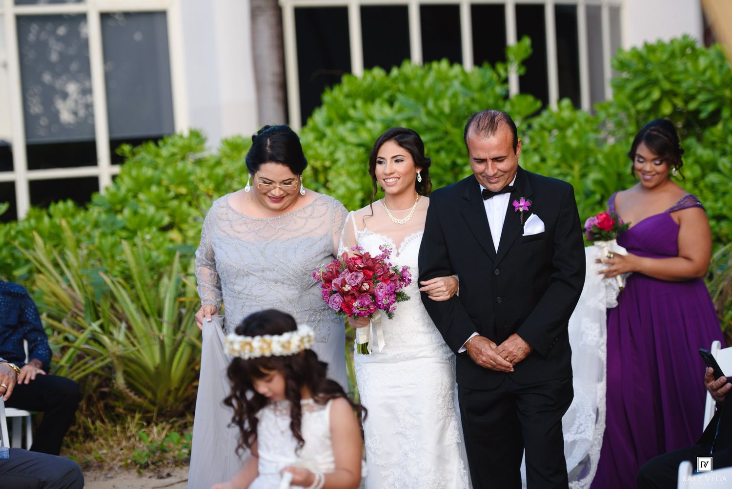 Bride walks down aisle with parents and flower girl at elegant outdoor wedding ceremony.