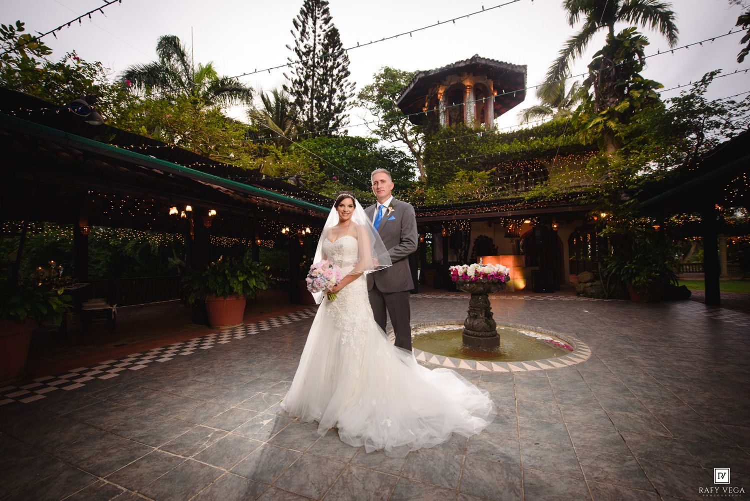 Bride and groom pose elegantly in a courtyard with string lights, fountain, and lush greenery at their outdoor wedding venue.