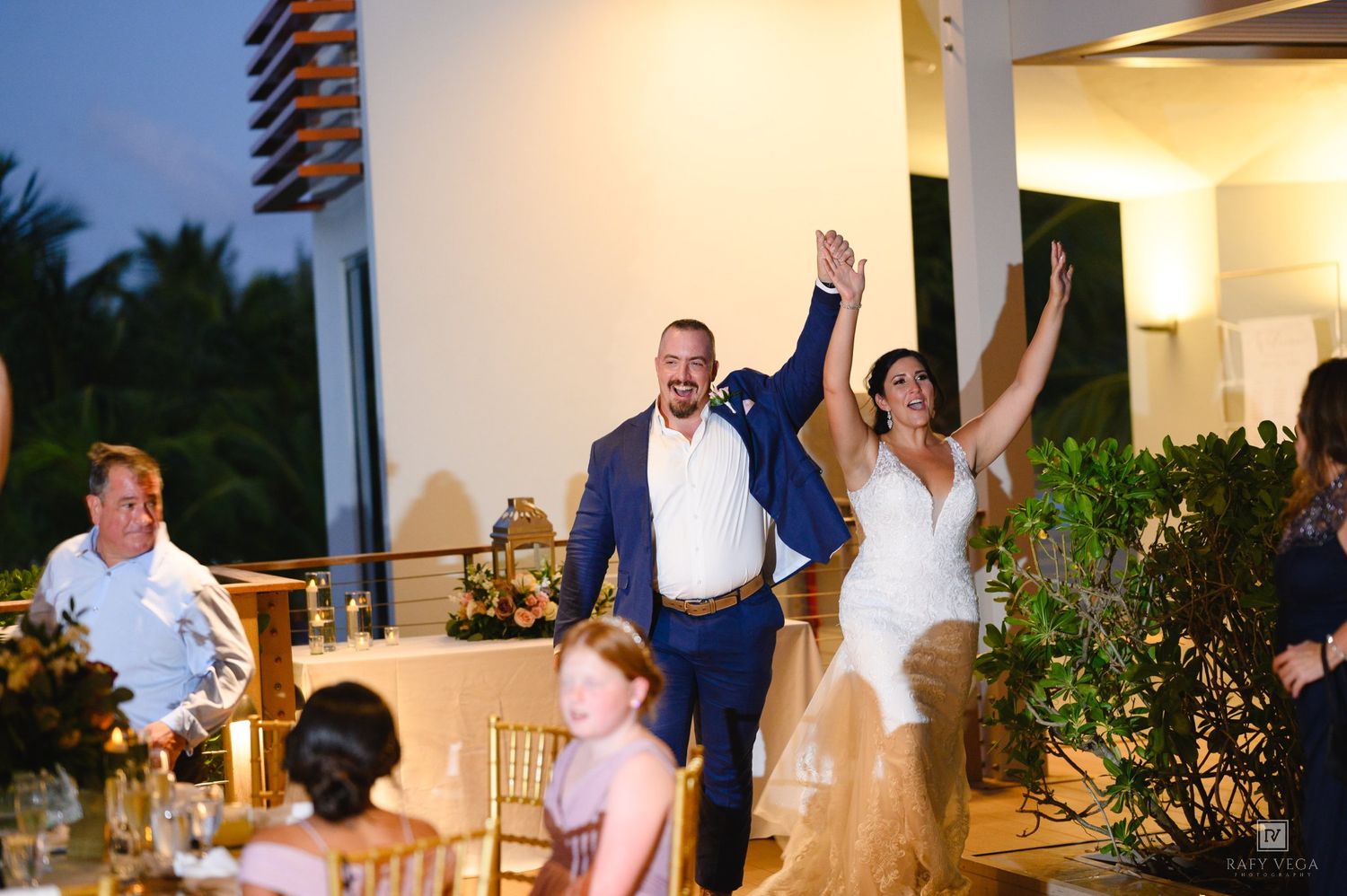 Bride and groom celebrate grand entrance at outdoor wedding reception, raising hands in joy among seated guests.