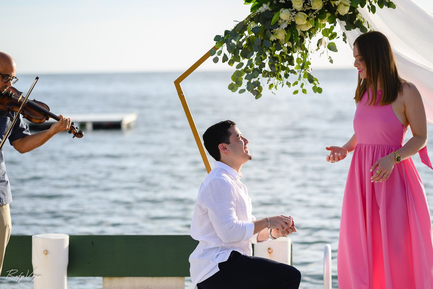 Man proposes on one knee to woman in pink dress by the sea, with violinist playing nearby under floral arch.