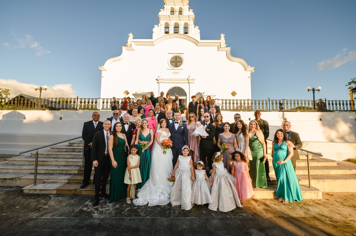 Large wedding party posing on steps outside white Spanish colonial church at sunset.