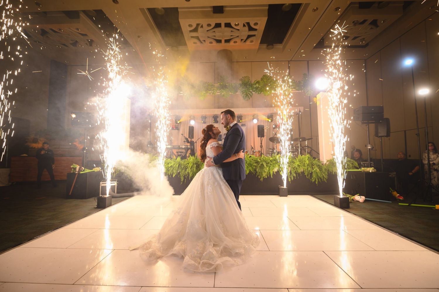 Bride and groom share first dance on white floor with dramatic sparkling fireworks fountain effects at elegant wedding reception.