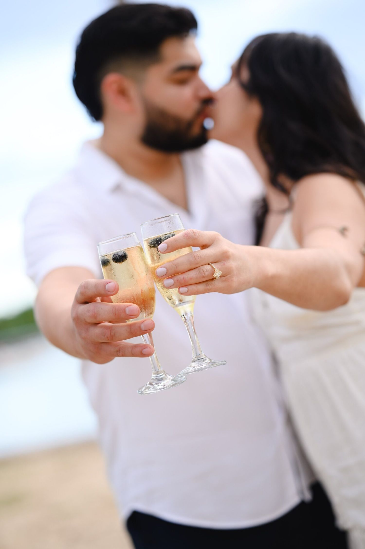 Engaged couple kisses while clinking champagne glasses on a beach during their proposal celebration.