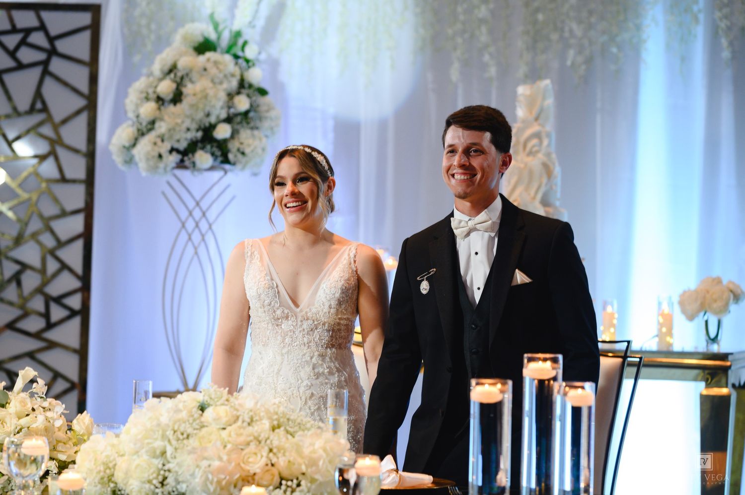 Bride and groom smiling at elegant white floral wedding reception with candles and decorative backdrop.