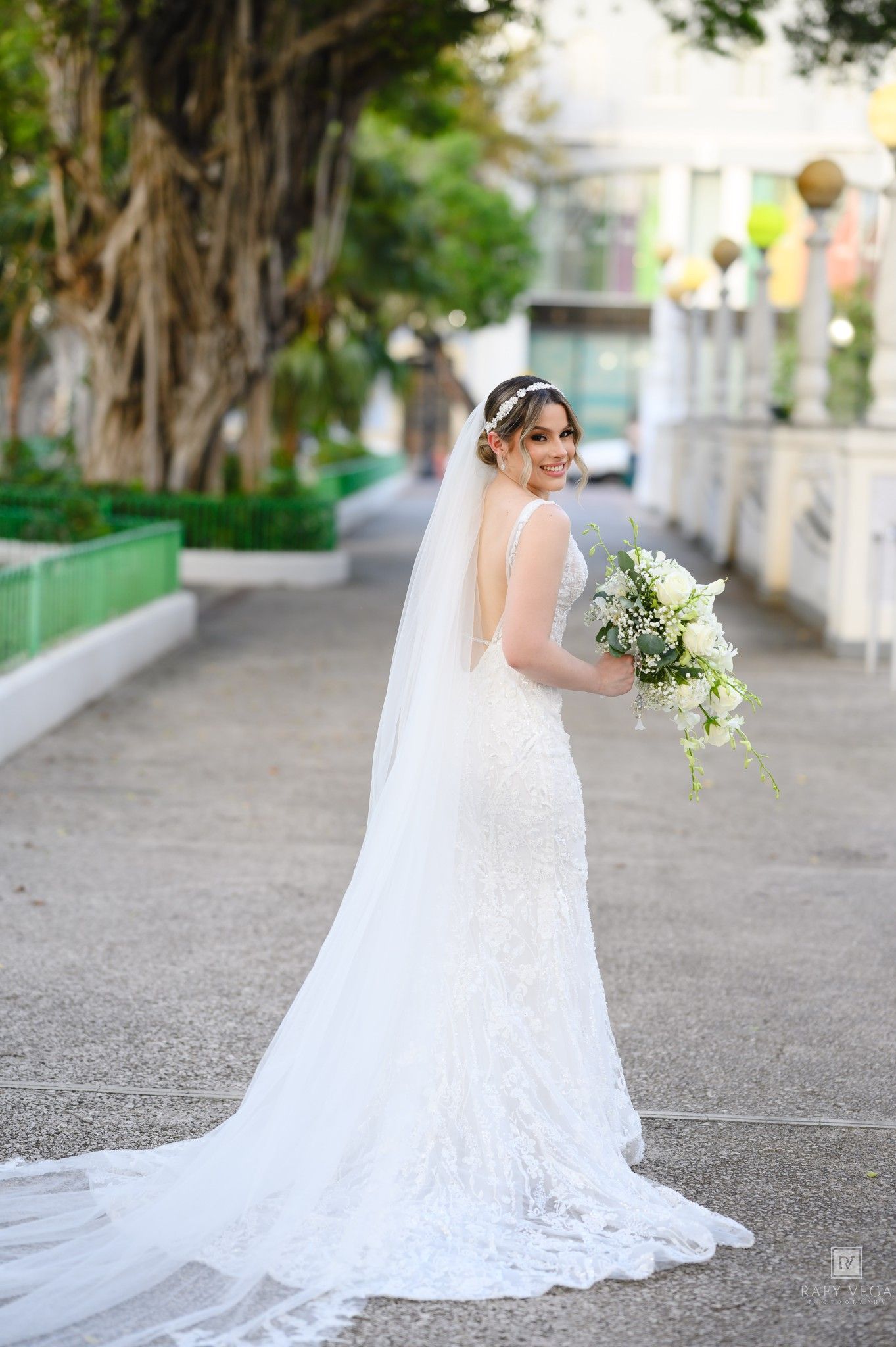 Bride in white lace gown with long veil holds bouquet, smiling over shoulder on outdoor path with trees.