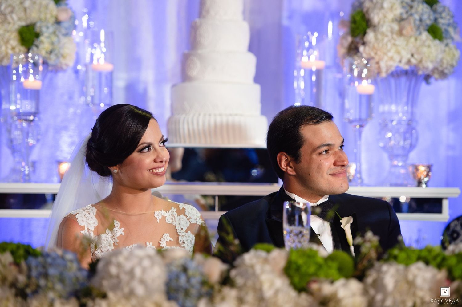 Bride and groom smiling at elegant wedding reception with white floral arrangements and tiered cake backdrop.