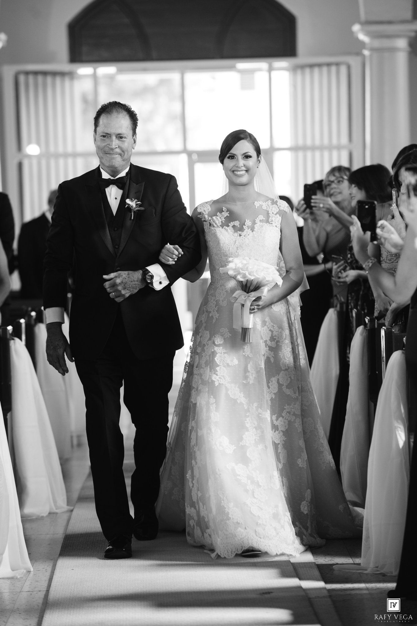 Bride in lace gown walks down aisle with escort in tuxedo during elegant wedding ceremony in black and white photo.