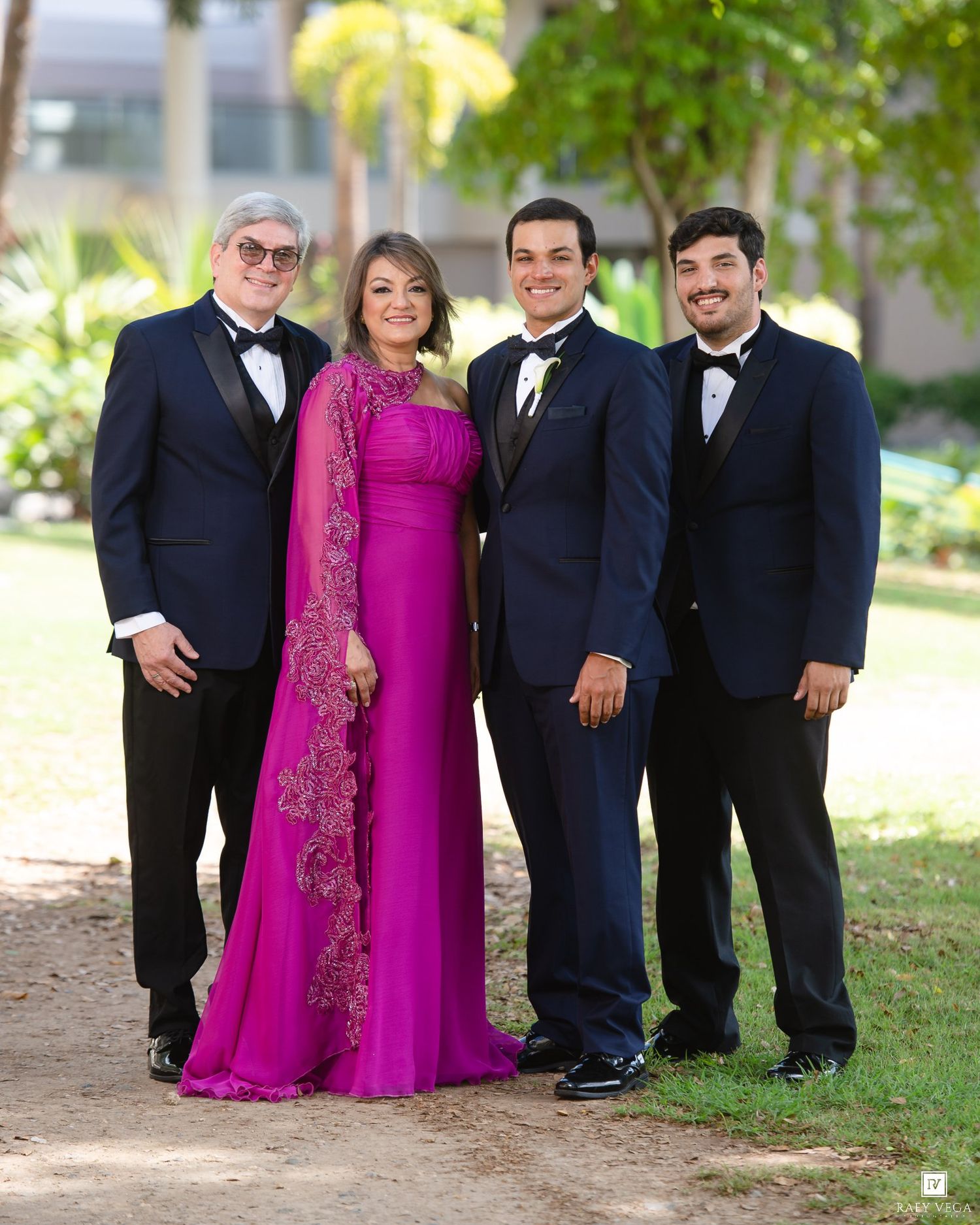 Family of four posing outdoors at a formal event, woman in elegant fuchsia gown, men in navy tuxedos.