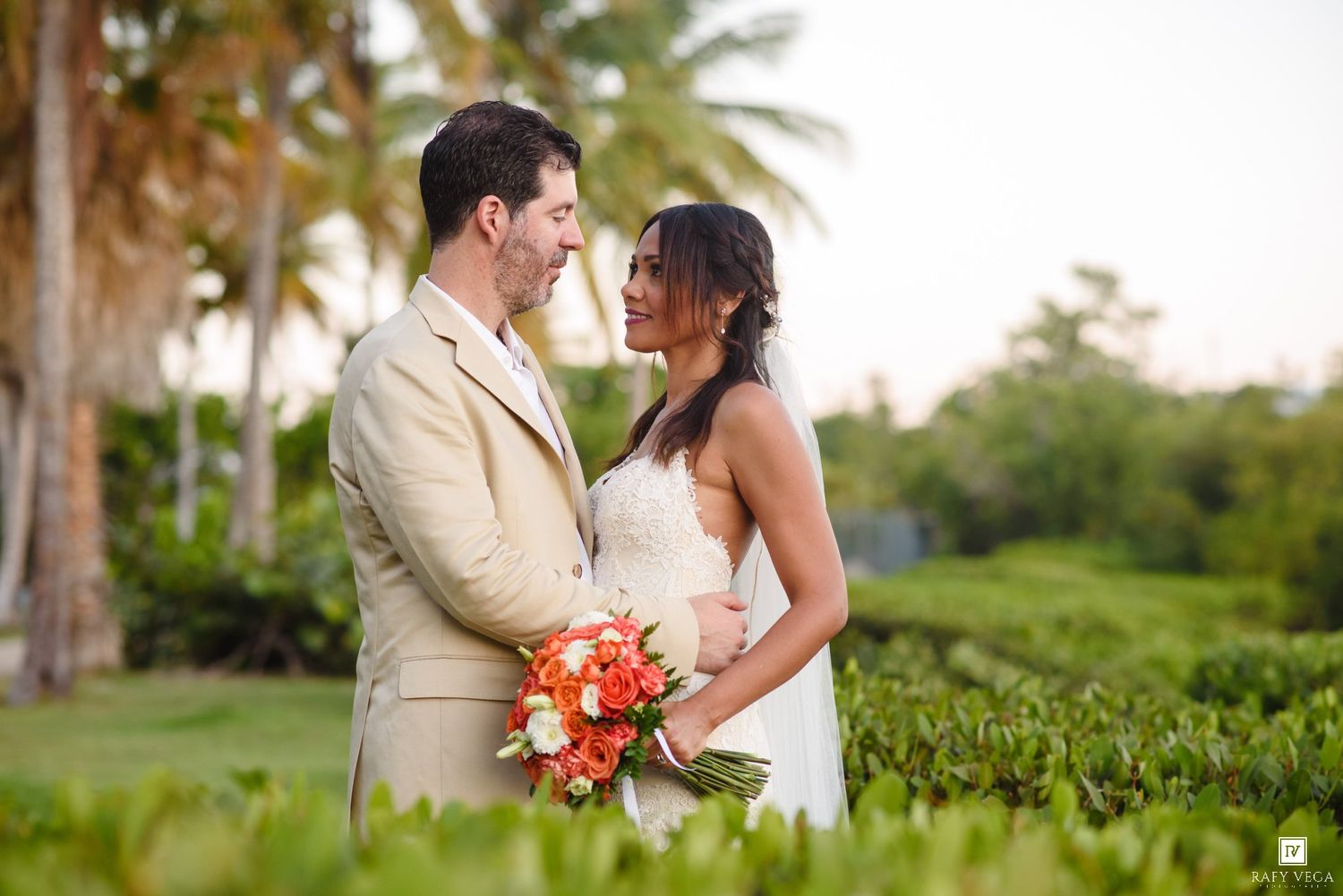 Bride and groom share a romantic moment outdoors, surrounded by tropical palms, as she holds a vibrant orange bouquet.