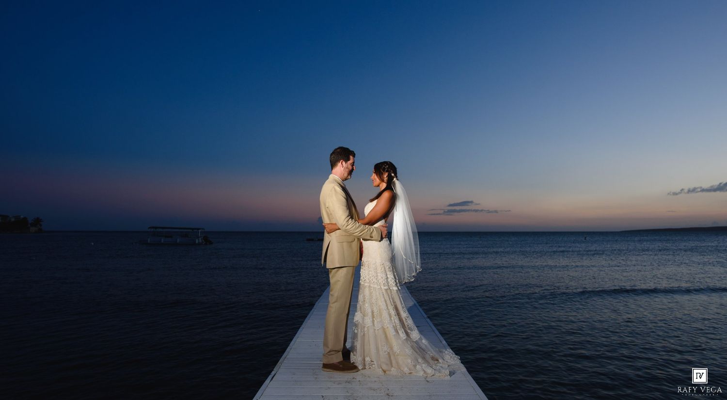 Couple shares romantic moment on pier at sunset over calm ocean, bride in white gown with veil, groom in tan suit.