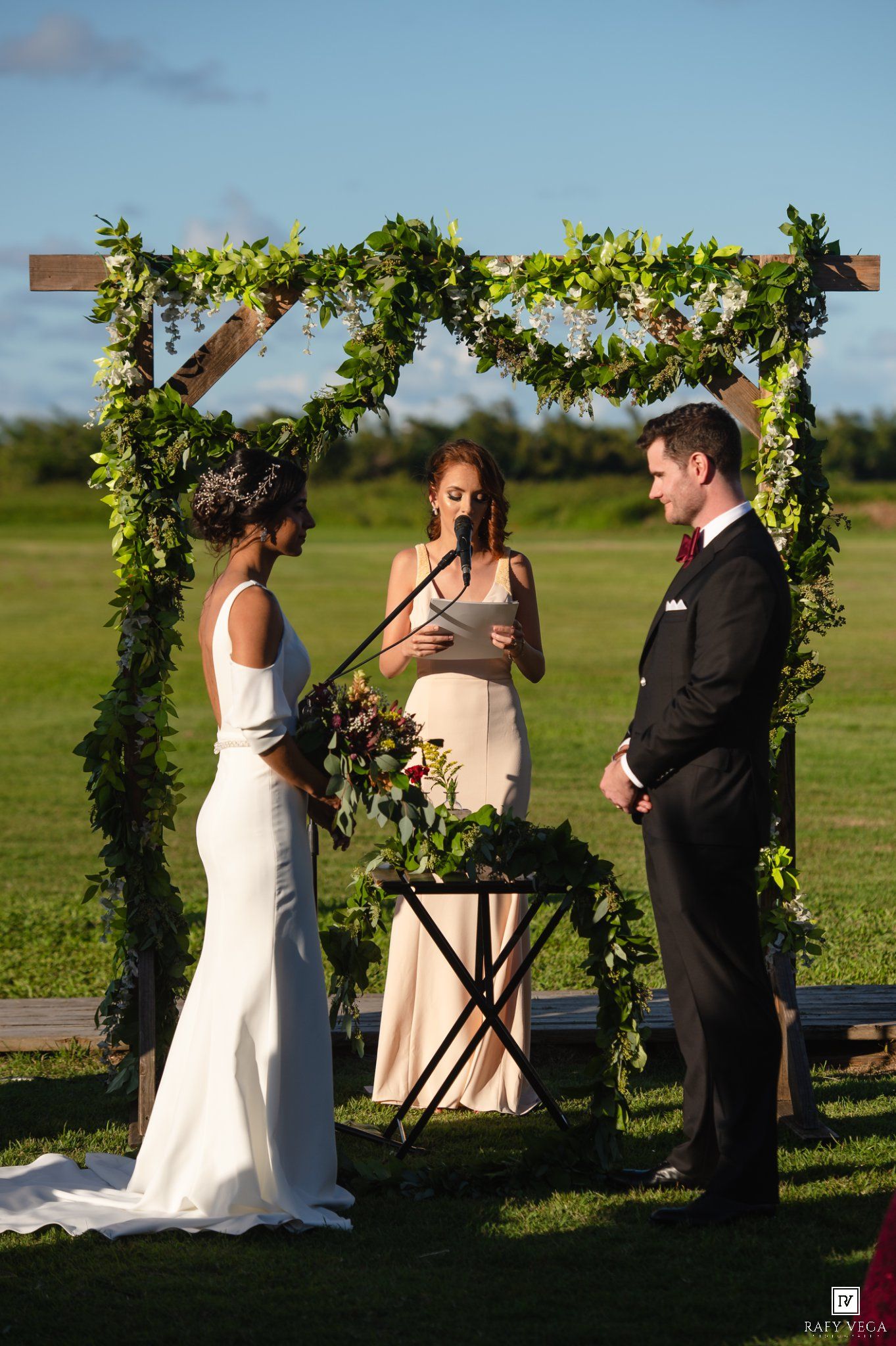 Outdoor wedding ceremony under a wooden arch adorned with lush greenery, bride in white, groom in black tuxedo.