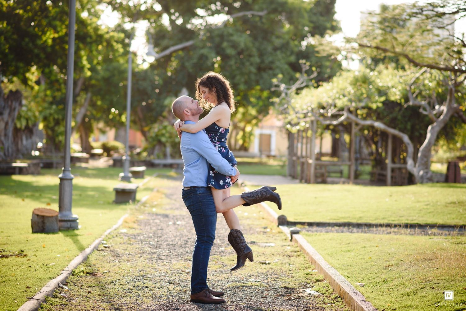 A couple shares a romantic moment in a sunny park, as the man lifts the woman joyfully on a tree-lined path.