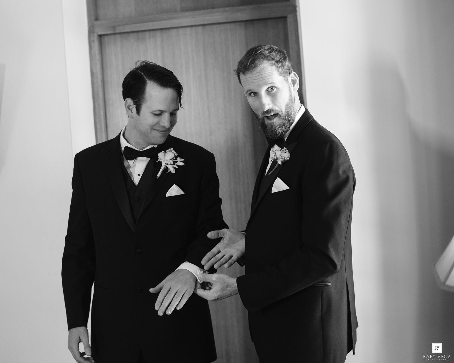 Two grooms in tuxedos admire wedding rings in a black and white photo on their wedding day.
