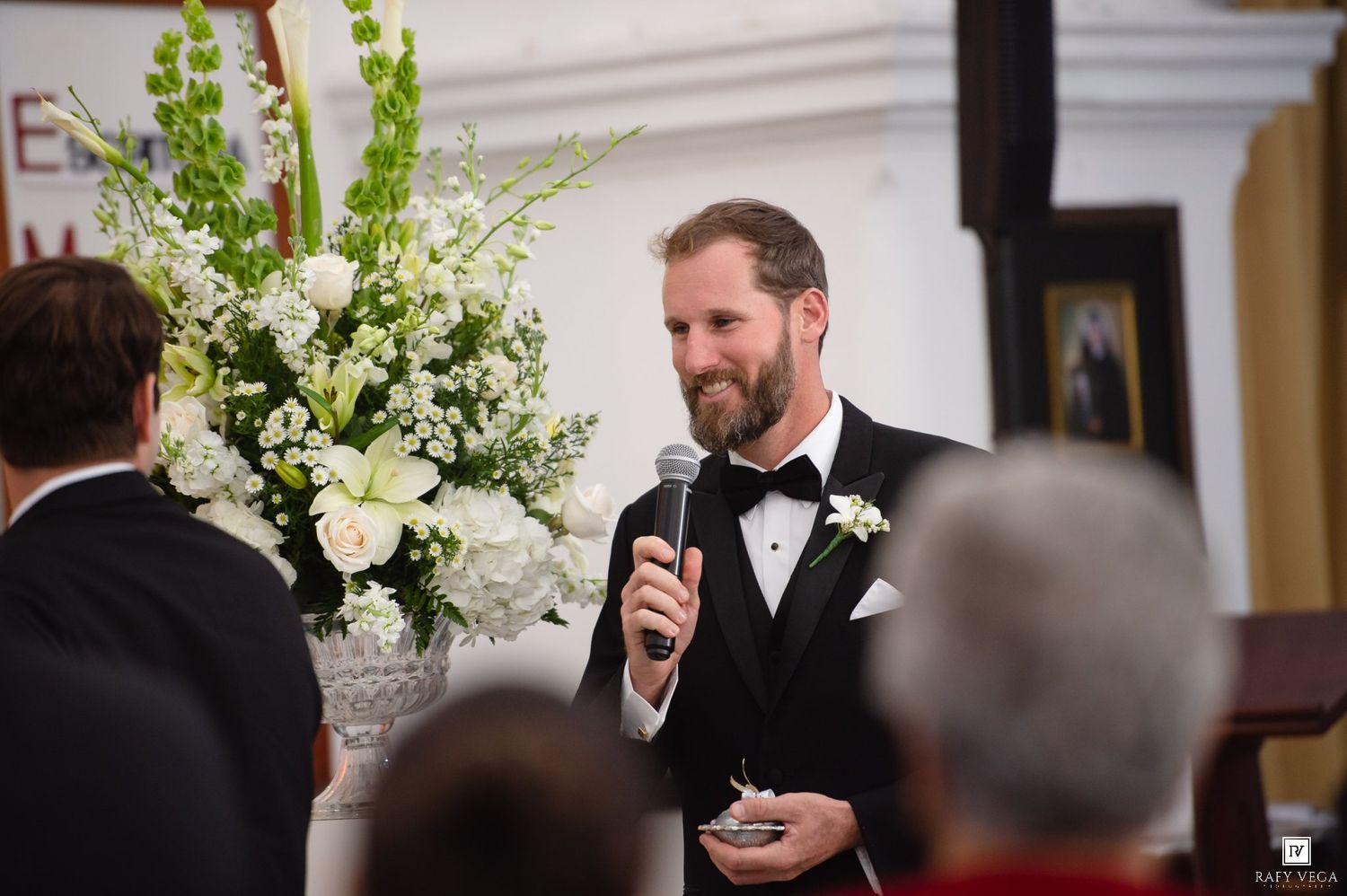 Bearded groom in tuxedo speaks into microphone at wedding ceremony near white floral arrangement.